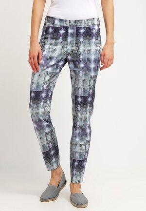 Kobieta nosząca dopasowane wzorzyste spodnie w odcieniach szarości, czerni i niebieskiego oraz szare buty wsuwane, stojąca na tle jednolitego tła.