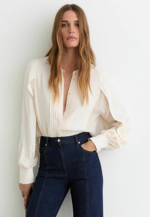 Femeie cu păr lung ondulat stă sprijinită de un perete alb, purtând o bluză plisată crem și blugi cu talie înaltă de culoare albastru închis, având o mână în buzunar.