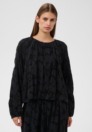 Donna con capelli castani lisci indossa una camicetta nera larga con motivo floreale e maniche lunghe su sfondo chiaro uniforme.