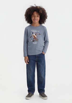 Otrok z kodrastimi lasmi, oblečen v sivo majico z dolgimi rokavi z grafiko pingvina na rolki, modre kavbojke in sive superge, stoji z roko v žepu.