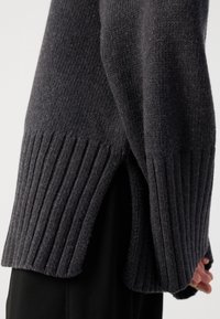 Pull en tricot gris foncé avec ourlet côtelé et fentes sur les côtés. Le tissu est doux et texturé, présentant une coupe ample qui complète sa structure.