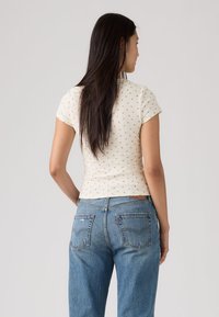 Γυναίκα με μακριά σκούρα μαλλιά που φοράει κοντομάνικο λευκό πουκάμισο με μικρά λουλουδάτα σχέδια και μπλε τζιν, στέκεται μπροστά σε απλό φόντο.