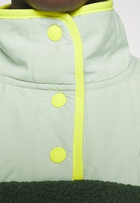 Jasnozielona kurtka z wysokim kołnierzem, kontrastującymi żółtymi wykończeniami i żółtymi guzikami. Posiada miękką, ciemnozieloną teksturę na dole.