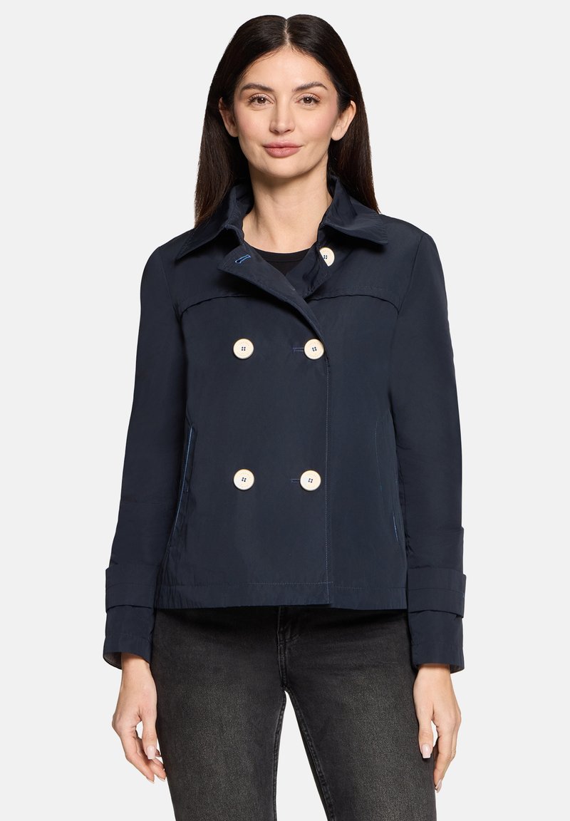 Vrouw draagt een marineblauw double-breasted jack met grote witte knopen en zwarte spijkerbroek, staand tegen een effen lichte achtergrond.