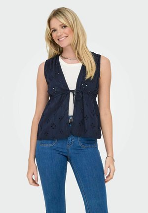 Top sin mangas color navy con detalles de ojal, escote en V, cierre de lazo en la cintura, usado sobre una camiseta blanca, combinado con jeans azules.