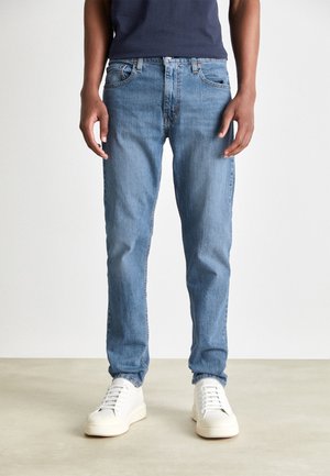 Homme portant un jean droit bleu clair et des baskets blanches, debout devant un fond clair uni.