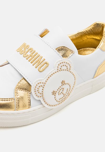 Sneaker bianca in pelle con accenti dorati, caratterizzata da un grande applicazione di orsetto di peluche e una fascia con logo in rilievo. Suola intermedia texturizzata e interno liscio.