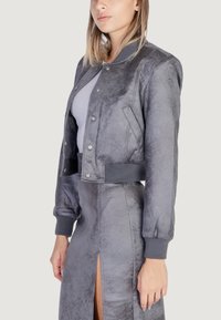 Chaqueta bomber gris con un acabado texturizado, botones a presión plateados, puños acanalados y cinturilla, combinada con una falda gris con una abertura lateral.