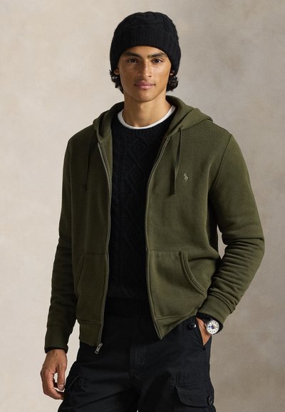 Olivgrüner Kapuzenpullover mit Reißverschluss und Vordertaschen, darüber ein dunkel strukturierter Pullover. Modell trägt eine schwarze Mütze und schwarze Cargohosen.