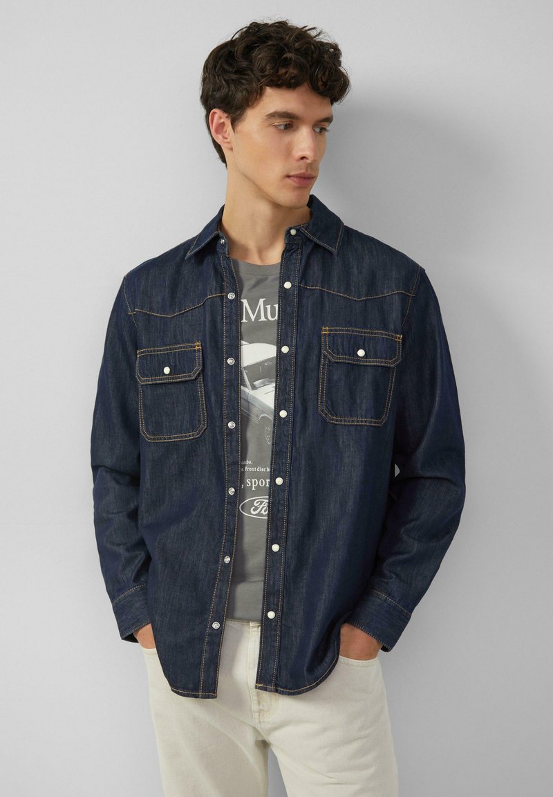 Donkerblauwe denim shirt met twee borstzakken, drukknoopsluiting en contrasterende oranje stiksels. Draag het over een grijs T-shirt met een opdruk.