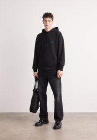 Schwarzer Kapuzenpullover mit blauem Logo, kombiniert mit schwarzen Jeans und schwarzen Schuhen. Das Modell hält eine schwarze Tragetasche und steht auf einer hellen Fläche.