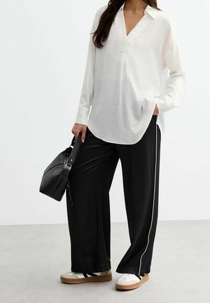 Vrouw die een losse witte blouse draagt, zwarte wijde broek met witte zijstrepen, witte en beige sneakers, en een zwarte handtas vasthoudt.