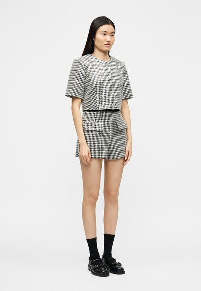 Femme debout portant une veste à manches courtes pied-de-poule noir et blanc et un short assorti, des chaussettes noires et des mocassins noirs à boucle sur fond blanc.