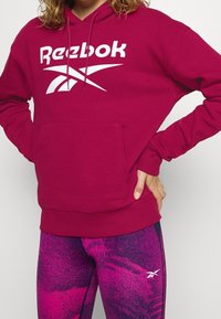 Marinröd huvtröja med en vit Reebok-logotyp, med en känguruficka och ribbade ärmslut. Kombination med rosa mönstrade leggings.