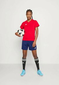 Punane sportlik särk V-kaelusega ja logoga, tumesinised lühikesed püksid, mustad põlvikud valgete triipudega, helesinised jalgpalliketsid, hoiab jalgpalli.