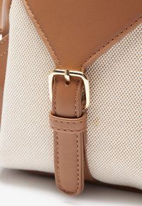 Correa de cuero marrón con herrajes dorados unida a una tela tejida en color crema, con una superficie texturizada y elementos de diseño angulares.