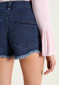 Džínové šortky s roztrhaným lemem v tmavě modré barvě, mají dvě zadní kapsy. Částečně viditelná je také světle růžová košile s dlouhým rukávem.