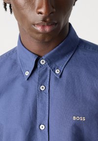 Marineblauwe knoopshirt van katoen, met een klassieke kraag, witte ronde knopen en een geborduurd 'BOSS'-logo in goud op de borst.
