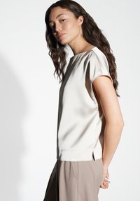 Top in seta beige chiaro con maniche corte; presenta una linea dritta e spacchi laterali per maggiore comfort. Il tessuto ha una consistenza liscia e morbida.