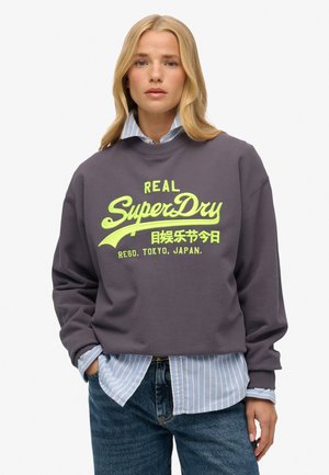 Donkergrijze sweatshirt met een groot, felgeel SuperDry-logo. Draagt over een blauw-wit gestreept shirt en gecombineerd met spijkerbroek.