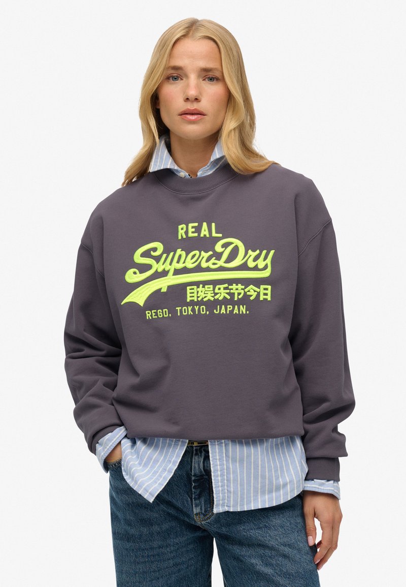 Donkergrijze sweatshirt met een groot, felgeel SuperDry-logo. Draagt over een blauw-wit gestreept shirt en gecombineerd met spijkerbroek.