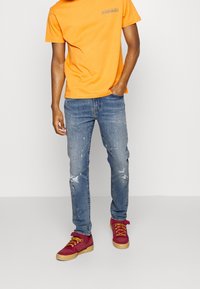 Orange bomull t-shirt, blå slitna jeans med hål, röda sneakers med gula snören och ljusbruna sulor.