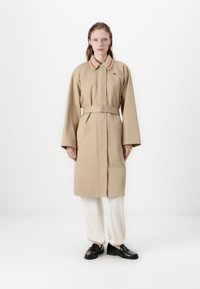 Trench coat beige con chiusura a bottoni, maniche ampie e vita aderente con cintura. Accenti notevoli di colore rosso e bianco sul colletto. Indossato su pantaloni chiari.