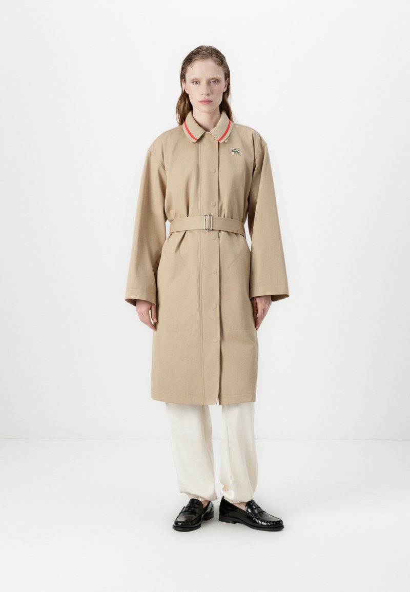 Trench coat beige con chiusura a bottoni, maniche ampie e vita aderente con cintura. Accenti notevoli di colore rosso e bianco sul colletto. Indossato su pantaloni chiari.