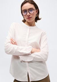 Γυναίκα που φοράει γυαλιά με μπλε απόχρωση, χρυσούς κρίκους στα αυτιά και λευκό μπλουζάκι με μανσέτες με βολάν, όρθια με σταυρωμένα τα χέρια.