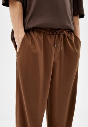 Osoba w brązowych spodniach ze ściągaczem i rękami w kieszeniach oraz w ciemnobrązowej koszuli na jednolitym tle.