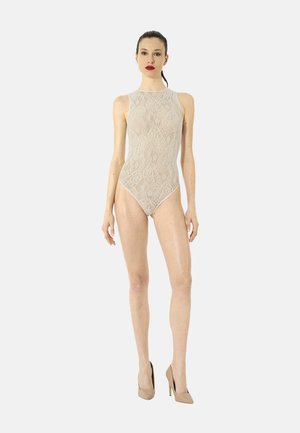 Donna in piedi che indossa un body in pizzo beige senza maniche e scarpe beige con tacco alto contro uno sfondo chiaro e semplice.