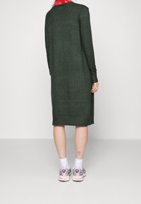 Mörkgrön långärmad klänning med rak skärning och ribbad fåll. Kombineras med grå sneakers och vita strumpor som avslöjar en tatuering på vristen.