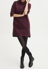 Bordowa sukienka z kołnierzykiem, długimi rękawami, zapięciem na guziki i zakrzywionym dołem, zestawiona z czarnymi legginsami i botkami za kostkę. Gładka tekstura materiału.