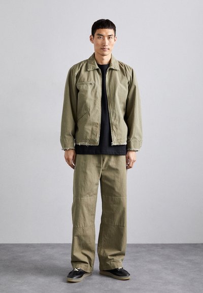 Olive green jacket with a zip front, two side pockets, and frayed hem, paired with matching loose-fit pants. Black t-shirt underneath, black shoes.
