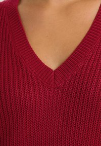 Roter Strickpullover mit tiefem V-Ausschnitt, gerippter Textur und sichtbaren Nähdetails. Weicher Stoff mit lockerer Passform.