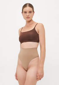 Donna con i capelli raccolti, che indossa un bralette marrone e un body beige a vita alta, su uno sfondo chiaro e semplice.