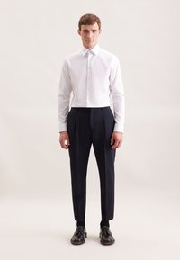 Man in een wit overhemd, zwarte pantalon, zwarte sokken en zwarte dresschoenen die tegen een effen lichte achtergrond staat.
