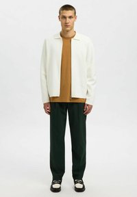 Jauns vīrietis stāv, ģērbies baltā ražotni zīmotāja jakā, brūnā krekļī, tumši zaļos biksēs un melnbaltās mokasīnās pret tukšu balto fonu.