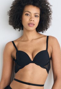 Soutien-gorge rembourré noir avec des accents en dentelle, doté de bretelles réglables, d'une surface lisse et d'un décolleté plongeant. Fermeture rectangulaire à l'arrière.