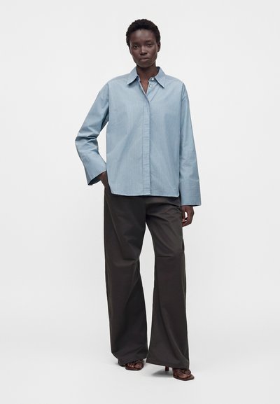 Femme debout portant une chemise ample bleu clair rayée à boutons, un pantalon large foncé et des sandales à talons sur un fond uni.