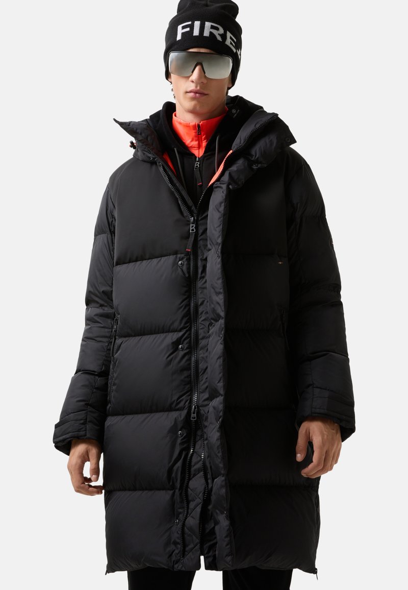 Person, der einen langen schwarzen Daunenmantel, eine schwarze Mütze mit dem Text "FIRE", eine reflektierende Sonnenbrille und eine orange-rote Zip-Up-Jacke darunter trägt.