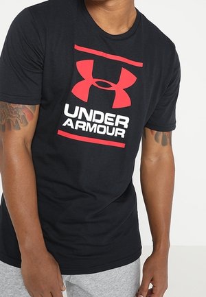 Osoba nosząca czarną koszulkę Under Armour z czerwonym logo i napisem, w połączeniu z jasnoszarymi spodniami, pokazująca tatuaże na ramionach.