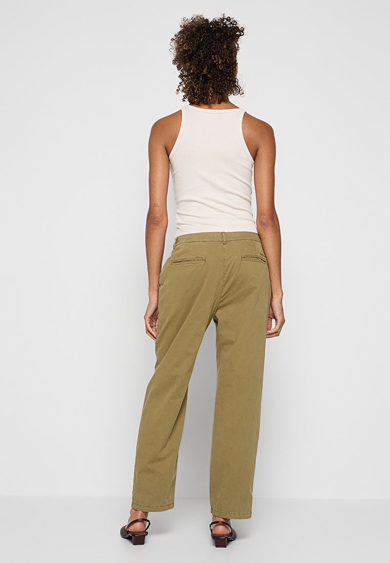 Frau mit lockigem Haar, die ein weißes ärmelloses Oberteil, olivgrüne weit geschnittene Hose und schwarze hohe Sandalen trägt, von hinten vor einem weißen Hintergrund blickend.