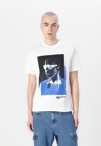 Bijela pamučna majica s grafičkim printom u crnoj i plavoj boji, prikazuje stilizirano lice, s natpisom "KARL LAGERFELD JEANS".