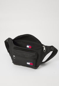 Schwarze Stoffbauchtasche mit zwei Reißverschlussfächern, ausgestattet mit einer kleinen Fronttasche und dem Tommy Jeans Logo in Rot, Weiß und Blau.