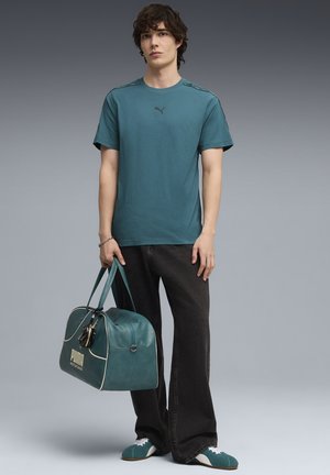 Młody mężczyzna w turkusowej koszulce i butach marki Puma, czarnych dżinsach, trzymający pasującą turkusową torbę sportową Puma na szarym tle.