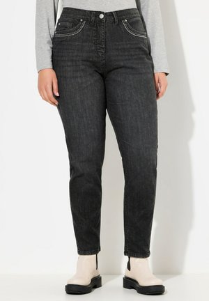 Frau trägt hoch taillierte schwarze Jeans mit dekorativen Nähten an den Taschen und hellbeige Stiefeletten, steht vor einem weißen Hintergrund.