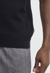 Pulover negru din tricot cu bordură, asortat cu pantaloni texturati gri deschis. Are un eticheta de brand vizibilă pe partea stângă.