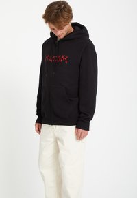Zwarte rits hoodie gemaakt van zachte stof met een voorzak en een rode grafische tekstontwerp. Gekocht met lichtgekleurde, loszittende broek.