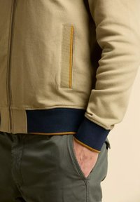Giacca a zip beige con tessuto testurizzato, polsini e orlo a costine blu navy, e una tasca accentuata con bordi gialli.